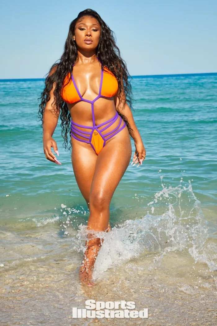 Megan Thee Stallion walks out of the water in an orange and purple string monokini.